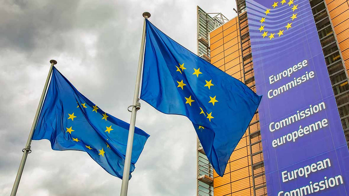 Gebäude mit der Aufschrift "Europöische Kommission", davor zwei EU-Flaggen. Im Hintergrund ein grauer Himmel. 