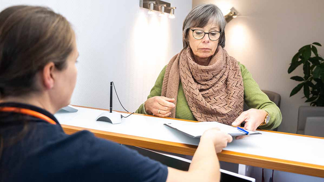 Eine Frau steht am Tresen einer Arztpraxis und erhält Unterlagen. 