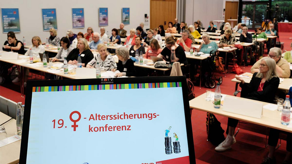 Ein Display mit dem Text "19. Frauenalterssicherungskonferenz", im Hintergrund de vollbesetzte Veranstaltungsraum. 