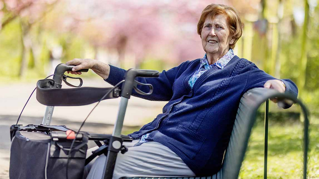 Ältere Frau mit Rollator sitzt auf einer Parkbank. 