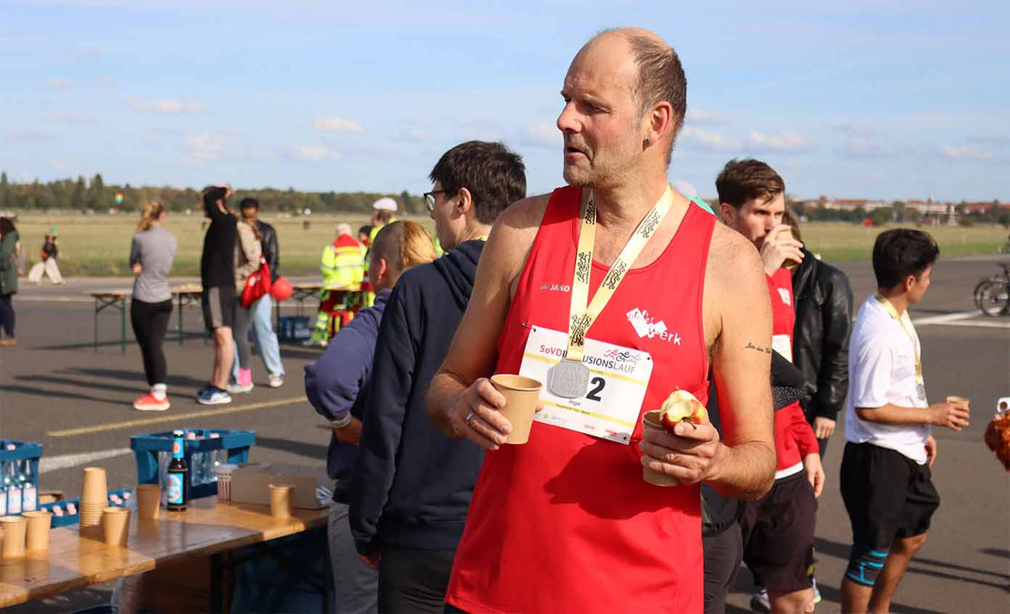 Mann mit Medaille um den Hals trinkt aus einem Becher und hat einen Apfel in der Hand. 